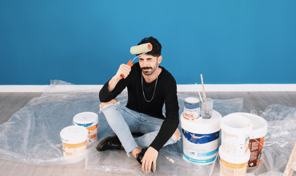 Painter before learning the steps to Become a Painting Business Pro.
Paint roller agains head grimacing while sitting on poly plastic surrounded by paint pails in a blue room.